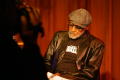 Photograph: [Photograph of an individual filming Melvin Van Peebles on stage]