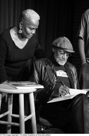 Primary view of object titled '[Photograph of a woman standing by Melvin Van Peebles as he signs books]'.