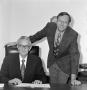 Photograph: [Two men smiling behind a desk]