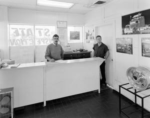 Primary view of object titled '[Two men standing behind a counter at a mechanic shop]'.