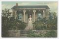 Postcard: [Postcard of an Older Woman Standing in Front of a House]