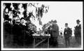 Photograph: [Group of men gathered around a table for a cook out at Camp George]
