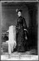 Primary view of [A woman standing beside a chair wearing a black dress and hat]