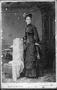 Photograph: [A woman standing beside a chair wearing a black dress and hat]