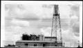 Primary view of [An oil derrick and a tin building with three storage tanks on top]